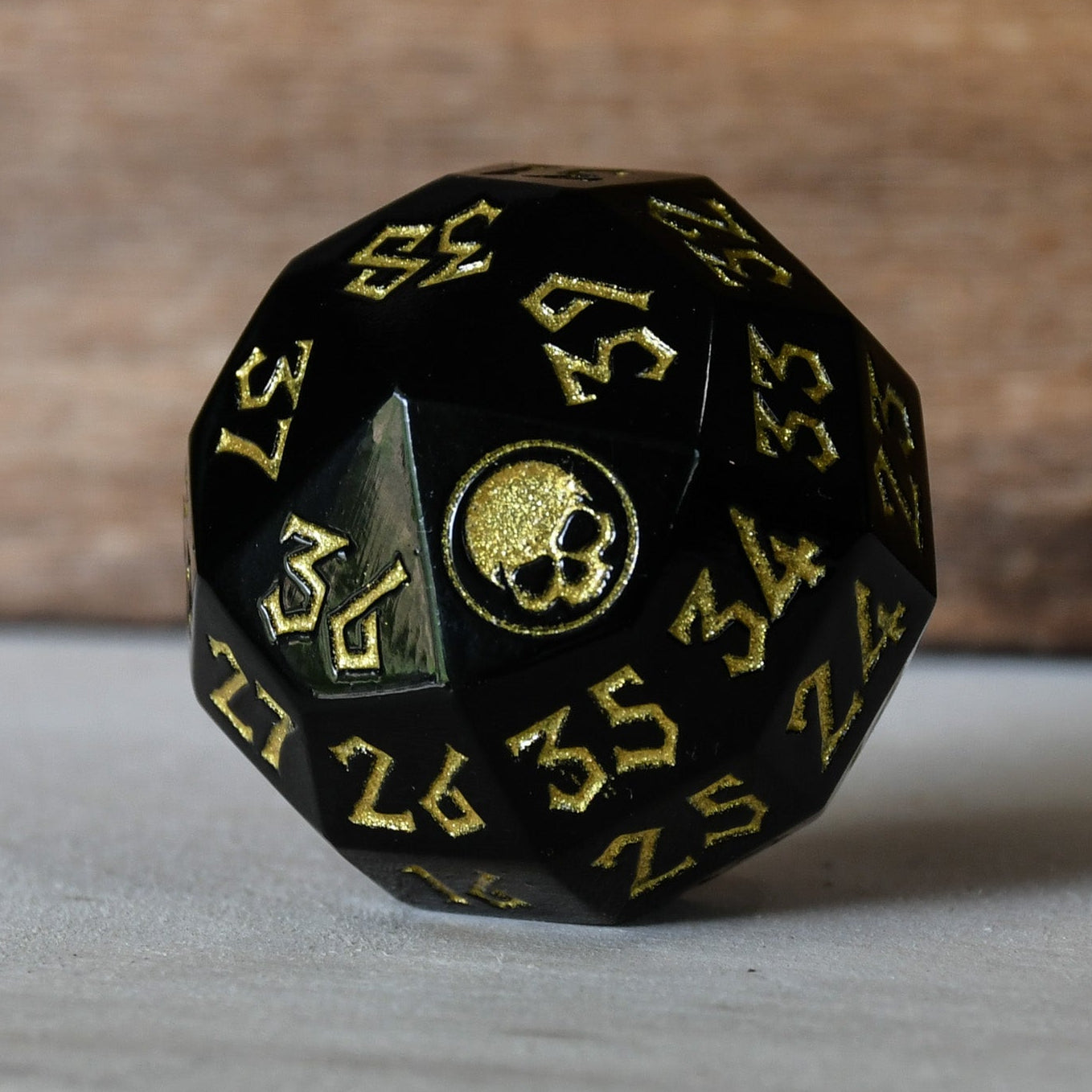 A solid-metal spindown life counter dice, black with gold numbers and iconography, displayed on a wooden surface.