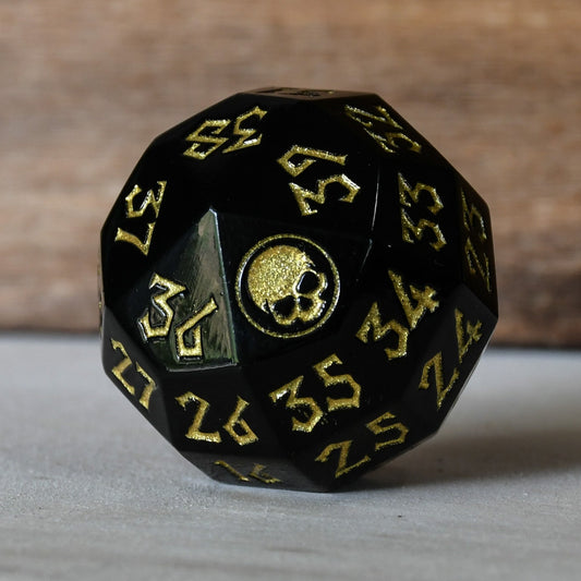 A solid-metal spindown life counter dice, black with gold numbers and iconography, displayed on a wooden surface.